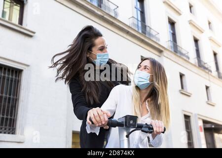 Due ragazze divertenti su uno scooter elettrico. Indossano maschere facciali. Concetto di nuova normalità.