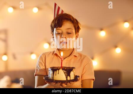 Ragazzo piccolo in un cappello da festa che soffia una candela sopra una piccola torta di compleanno e fare un desiderio Foto Stock