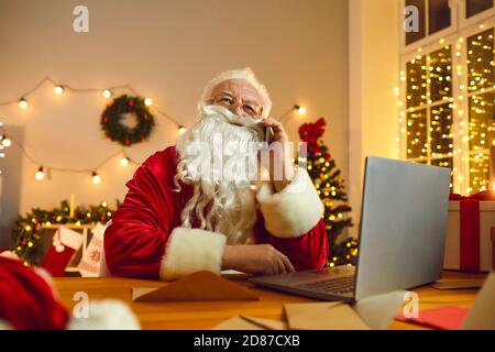 Babbo Natale lavora su un computer portatile mentre parla per telefono in camera decorata per Natale. Foto Stock