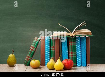 Libro di testo aperto, mucchio di libri in copertine colorate, pere e mele su tavola di legno con sfondo verde lavagna. Istruzione domestica di distanza. Torna a s Foto Stock