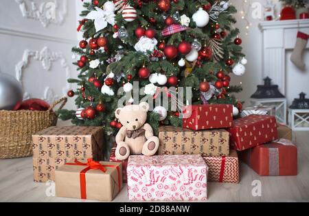 Regali in scatole e un orsacchiotto da vicino sotto un albero di Natale con decorazioni rosse e bianche in un soggiorno bianco. Anno nuovo, stile europeo, cucina dic Foto Stock