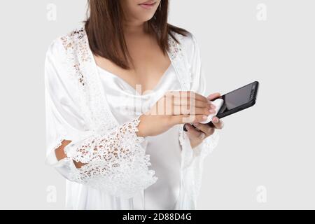 Una donna in bianco da notte che pulisce il suo telefono cellulare sullo schermo con il cotone su uno sfondo grigio chiaro, pulizia smartphone per eliminare i germi. Foto Stock