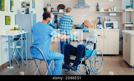 Geriatriciano che dà pillole a pazienti anziani disabili in sedia a rotelle durante la visita casa del coronavirus. Infermiera maschile lavoratore sociale in coppia anziani disabili con disabilità spiegare la diffusione del covid-19 Foto Stock