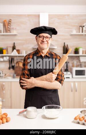 Vecchio uomo caucasico che indossa grembiule in cucina casalinga sorridendo alla macchina fotografica. Panettiere anziani in cucina uniforme preparare ingredienti di pasticceria su tavola di legno pronti a cucinare pane, torte e pasta gustosi fatti in casa Foto Stock