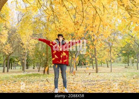 giovane donna con capelli ricci neri in pullover rosso e. il cappello di lana ha divertimento gettando le foglie asciutte che sono caduti dagli alberi in un parco in autunno Foto Stock