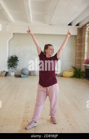 Giovane donna in abbigliamento sportivo alzando le mani e sorridendo si esercita in studio di danza Foto Stock