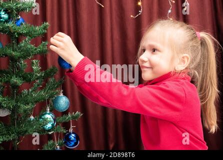 Una ragazza allegra caucasica carina in un maglione rosso appende una palla di festa su un albero di Natale artificiale decorato, un bambino decora un albero di Natale. HAP Foto Stock