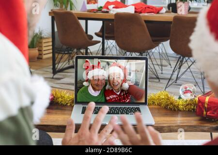 Vista posteriore di uomo e figlio in santa cappelli sventolando mentre si ha una videocall con la coppia senior a santa h. Foto Stock