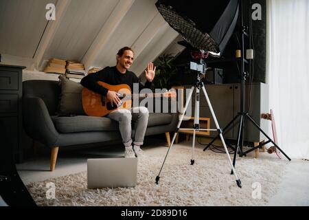 Musicista maschio caucasico che chiacchiera in videochiamata con la band che suona chitarra in una comoda camera da letto Foto Stock
