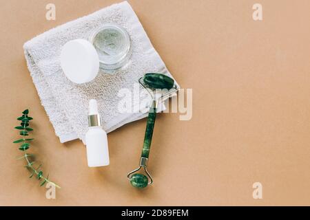 Siero cosmetico in flacone bianco senza marchio, vasetto crema e rullo facciale. Asciugamano, foglie di eucalipto su sfondo beige. Vista dall'alto, disposizione piatta, spazio di copia. Foto Stock