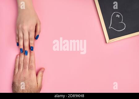 Mano di donna sconosciuta sta toccando uno mans uno contro sfondo rosa, accanto a una piccola lavagna con cuore disegnato su di esso. Primo piano, spazio di copia Foto Stock