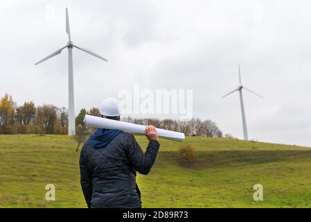 Vista posteriore di un ingegnere maschio indossa il casco bianco che regge un piano a turbina eolica sito autunno nuvoloso cielo verde prato Foto Stock