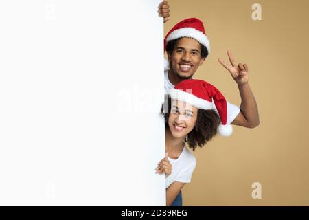 Sorridente giovane coppia afroamericana in cappelli di Natale che sbirciano fuori da dietro il banner bianco vuoto Foto Stock