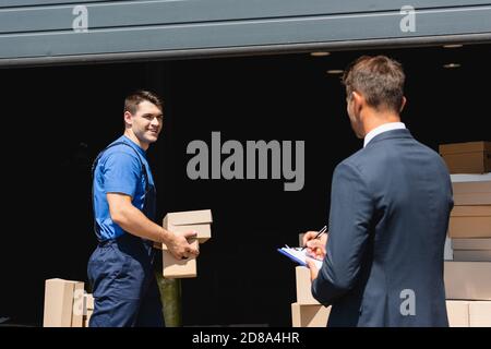 Fuoco selettivo del caricatore che tiene le scatole di cartone vicino alla scrittura del uomo d'affari su portadocumenti e magazzini all'aperto Foto Stock