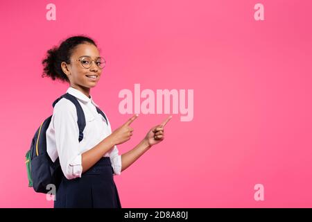 sorridente studentessa afroamericana in occhiali con zaino che punta da parte isolato su rosa Foto Stock