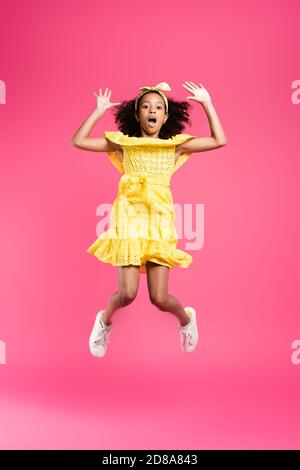 visione completa di un bambino afro-americano scioccato e curly outfit giallo che salta su sfondo rosa Foto Stock