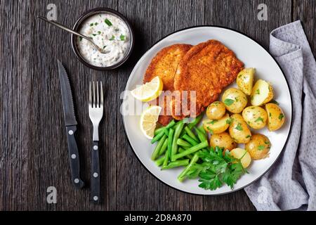 Schnitzel di maiale tedesco con patate giovani, fagioli verdi con posate servite su un piatto con spicchi di limone, e salsa a base di maionese su una b di legno scuro Foto Stock