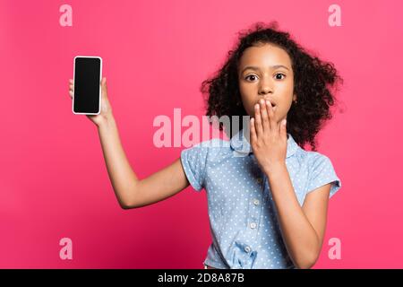 scioccato capretto afro-americano che presenta smartphone isolato su rosa Foto Stock