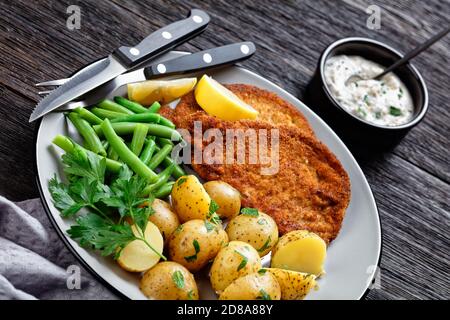 Schnitzel di maiale tedesco con patate giovani, fagioli verdi con posate servite su un piatto con spicchi di limone, e salsa a base di maionese su una b di legno scuro Foto Stock