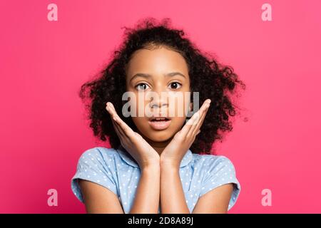 scioccato carino ricciolo afroamericano con le mani vicino al viso isolato su rosa Foto Stock