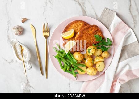 Schnitzel di maiale impanato con patate giovani, fagioli verdi con posate e salsa maionese serviti su un piatto rosa con spicchi di limone, su un marmo chiaro Foto Stock