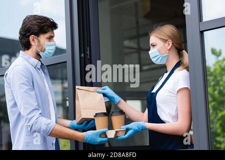 Vista laterale dell'uomo con guanti in lattice e maschera medica ricezione dell'ordine dalla cameriera vicino al bar Foto Stock