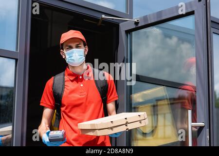 Fuoco selettivo del corriere nella maschera medica che tiene il terminale di pagamento e pizzerie vicino all'edificio sulla strada urbana Foto Stock