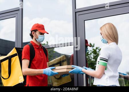 Corriere in maschera medica e guanti in lattice che danno scatole per pizza a donna vicino a costruire su strada urbana Foto Stock