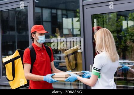 Uomo di consegna in maschera medica che dà scatole di pizza a donna in guanti di lattice su strada urbana Foto Stock