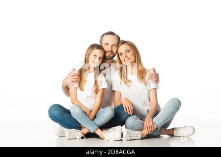 felice famiglia seduta sul pavimento con gambe incrociate e abbracciando isolato su bianco Foto Stock