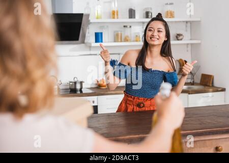 fuoco selettivo della donna vicino amico asiatico eccitato che mostra i pollici in cucina Foto Stock
