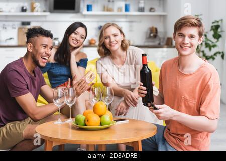 giovane che tiene una bottiglia di vino rosso vicino ad amici multiculturali e frutta fresca in cucina Foto Stock