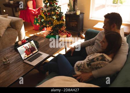 Coppia seduta sul divano con una videocall con coppia in babbo natale sorride su un computer portatile a casa Foto Stock
