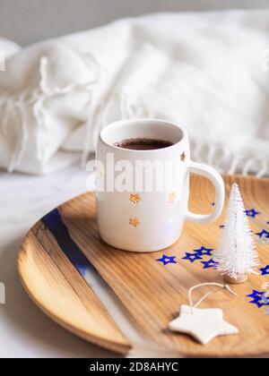 Tazza in ceramica con stelle con caffè nero, albero di natale bianco e stella tramica su un vassoio su sfondo chiaro. Stile di vita. Vista frontale. Foto Stock