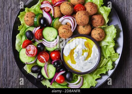 Piatto mediterraneo di falafel, pomodori, cetrioli, cipolla rossa, olive nere, sulle foglie fresche di insalata servite con salsa tzatziki servita su un nero Foto Stock