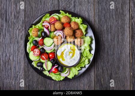 Piatto mediterraneo di falafel, pomodori, cetrioli, cipolla rossa, olive nere, sulle foglie fresche di insalata servite con salsa tzatziki servita su un nero Foto Stock