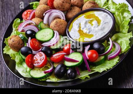 Piatto mediterraneo di falafel, pomodori, cetrioli, cipolla rossa, olive nere, sulle foglie fresche di insalata servite con salsa tzatziki servita su un nero Foto Stock