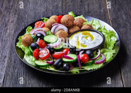 Piatto mediterraneo di falafel, pomodori, cetrioli, cipolla rossa, olive nere, sulle foglie fresche di insalata servite con salsa tzatziki servita su un nero Foto Stock