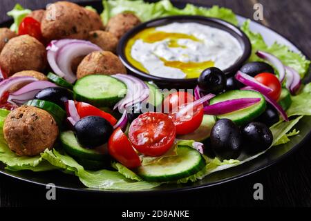 Piatto mediterraneo di falafel, pomodori, cetrioli, cipolla rossa, olive nere, sulle foglie fresche di insalata servite con salsa tzatziki servita su un nero Foto Stock