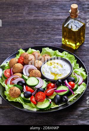 Piatto mediterraneo di falafel, pomodori, cetrioli, cipolla rossa, olive nere, sulle foglie fresche di insalata servite con salsa tzatziki servita su un nero Foto Stock