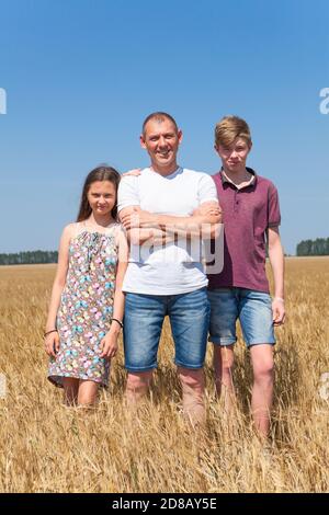 Papà con figlio preseato e figlia, ritratto a lunghezza intera, padre singolo con due bambini in piedi su campo di grano in estate Foto Stock