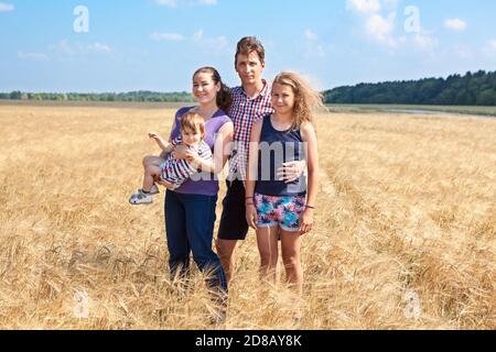 Giovane famiglia con due figlie di diverse età, ragazze preseine e infantili, quattro persone in piedi su campo di grano giallo, ritratto a lunghezza intera Foto Stock