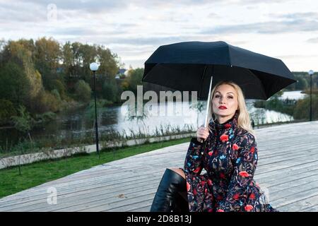 Una donna con ombrello nero si siede su una scala di legno, sullo sfondo di nuvole sul palco, con un bel sorriso Foto Stock