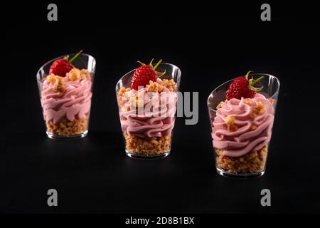 Vista in primo piano del dolce fresco fatto in casa in tre piccoli bicchieri di fila isolati su sfondo nero. Gustoso parfait con muesli, decorato con lamponi e panna montata rosa. Foto Stock