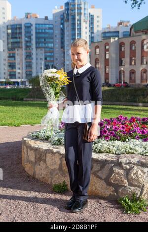 Ritratto a lunghezza intera della studentessa russa che indossa l'uniforme da scuola in piedi con fiori all'aperto, sorridente e guardando la macchina fotografica Foto Stock