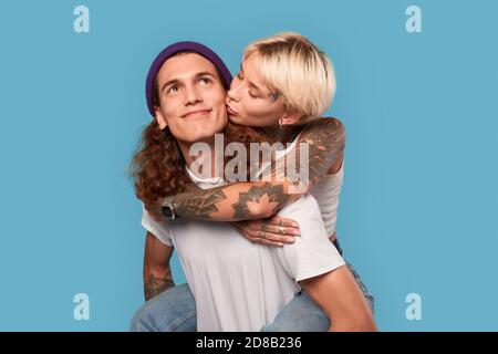 Stile libero. Giovane coppia tatuata che si trova isolata sulla ragazza blu che cavalca la schiena del ragazzo baciandolo mentre guarda da vicino sognante Foto Stock
