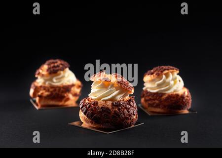 Tre deliziose palle di profiterole fresche e croccanti in fila con crema bianca dolce all'interno. Primo piano di gustosi eclari marroni fatti in casa isolati su sfondo nero. Concetto di dessert, cibo da ristorante. Foto Stock