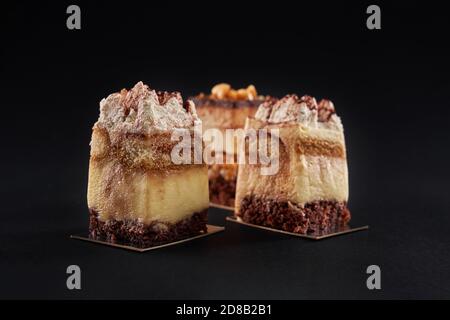 Vista in primo piano di tre pezzi quadrati in fila decorati con panna montata e cacao, isolati su nero. Torta tiramisù con tre strati di biscotto al cioccolato e sciroppo di caffè con cognac e crema. Foto Stock