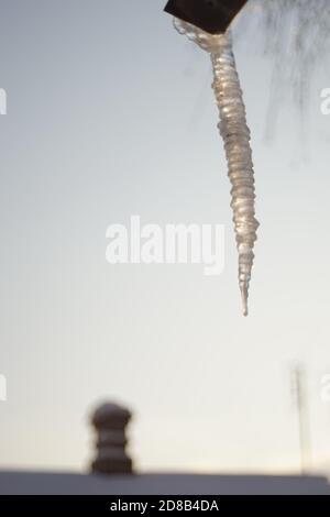 Enorme lunga icicola appesa su un tubo di drenaggio di una casa. Sfondo cielo blu e tetto rurale con camino. Foto Stock