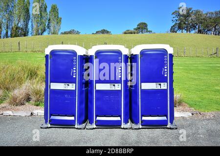 Fila di tre bagni portatili in plastica blu su ghiaia di fronte al pascoli verde recintato. Foto Stock